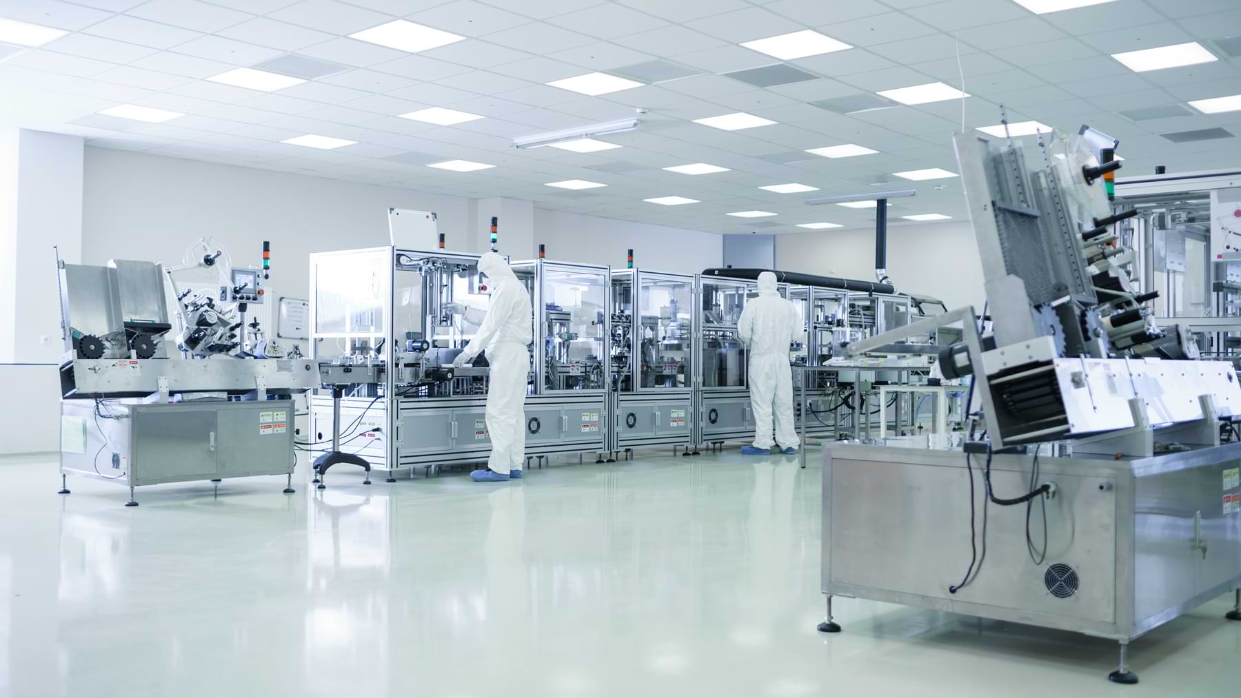 Zwei Arbeiter in Schutzanzügen bedienen moderne Maschinen in einer sterilen Produktionsstätte mit heller Beleuchtung und sauberen Böden.